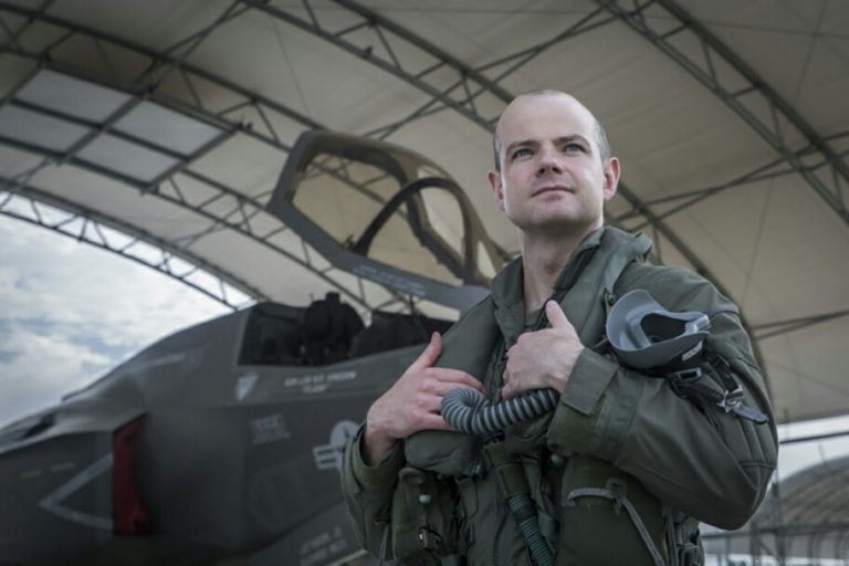 The veteran pilot, clad in a flight suit, stands confidently in front of an aircraft under a hangar, holding his helmet.