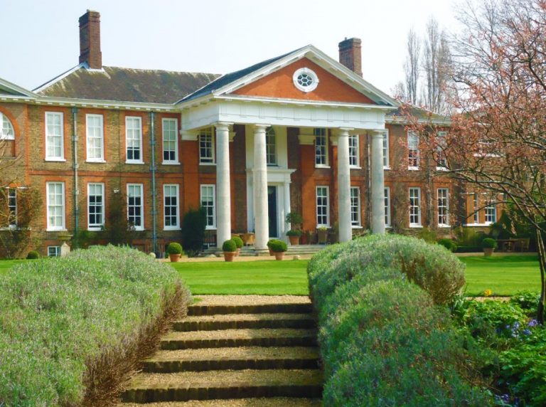 A large, classic brick mansion with white columns and a landscaped garden stands proudly, reminiscent of a veteran's resettlement home, as viewed from the stone steps leading up to it.