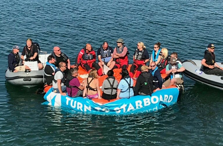 A group of veterans wearing life jackets are sitting on an inflatable raft with "Turn to Starboard" written on it, floating in the water.