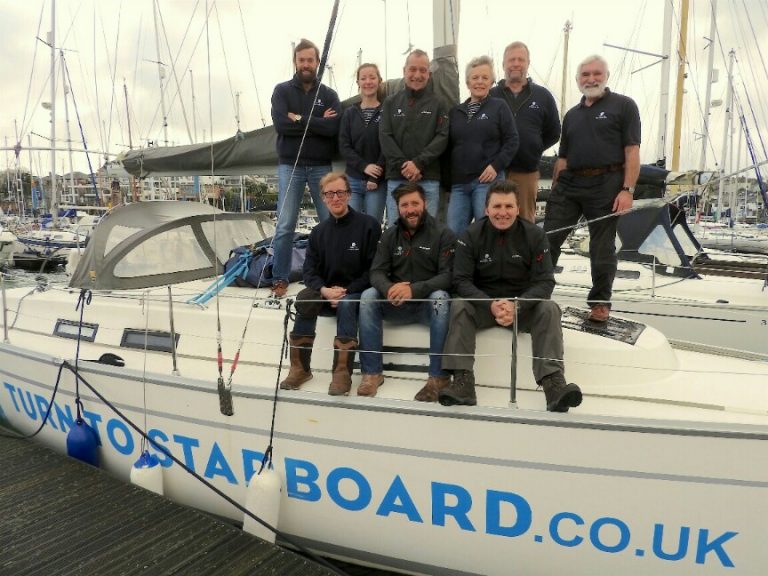A group of nine veterans in matching dark jackets pose together on a sailboat docked in the marina. With "turn to starboard.co.uk" written on the side, their camaraderie reflects their shared experiences, possibly as Royal Navy pathfinders, united by the sea.