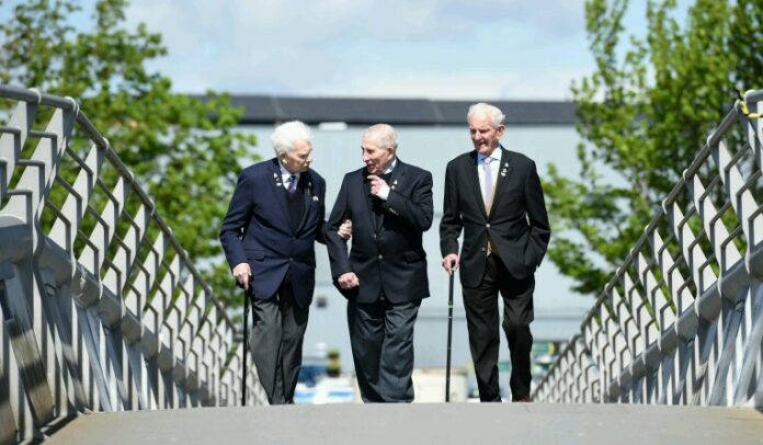 Three elderly men in suits, with medals on their lapels, likely veterans from the Royal Navy, are walking on a bridge. Two use canes. Trees and a building are visible in the background.