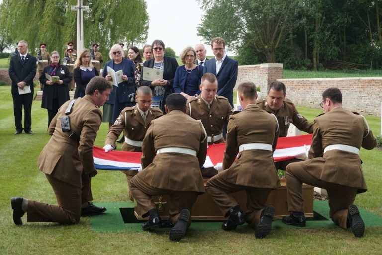 Military personnel in uniform solemnly lower a flag-draped casket at a graveside ceremony, honoring a respected veteran, surrounded by onlookers in the cemetery.