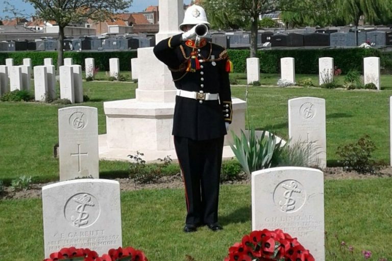 A uniformed bugler, a proud Royal Navy veteran, plays at a military cemetery, standing among white headstones with a white monument in the background, surrounded by greenery and wreaths.