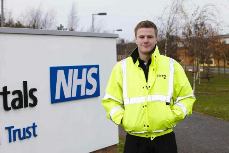 A person in a bright yellow jacket stands next to an NHS sign outside a building, reminiscent of the unity seen in the Royal Navy.
