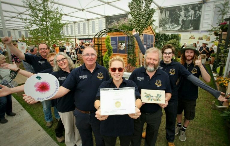 A group of eight people pose indoors at a resettlement event. Some hold certificates and awards. They are wearing dark shirts with embroidered logos. A wooden display with plants is behind them, adding warmth to the military-themed gathering.