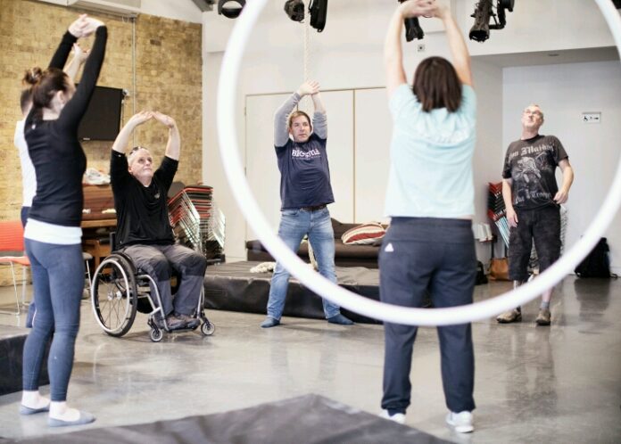 In a circle-shaped room setup, a diverse group of people, including a Royal Navy veteran in a wheelchair, stretch their arms in unison.