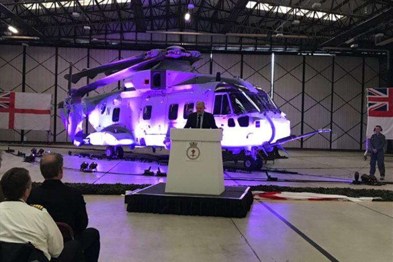 A veteran gives a speech at a podium in front of a helicopter inside a hangar, with Royal Navy flags displayed on the walls.