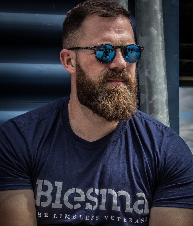 Bearded man wearing sunglasses and a "Blesma: The Limbless Veterans" t-shirt sits outdoors, embodying the spirit of a proud military veteran.