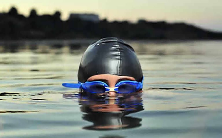 A swimmer, resembling a Royal Navy pathfinder, wearing a black swim cap and blue goggles, emerges from calm water at dusk.