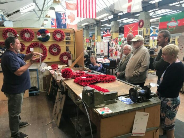 A group of people listens to a presentation in a workshop filled with poppy wreaths, flags, and various army memorabilia.