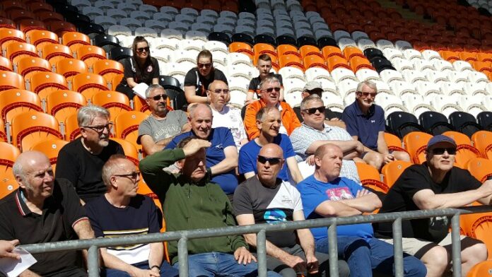 A group of people, including a few in Royal Navy attire, are sitting in a stadium with orange and white seats on a sunny day.