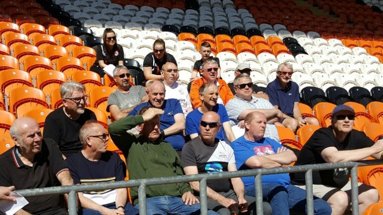 A group of people, including a few in Royal Navy attire, are sitting in a stadium with orange and white seats on a sunny day.