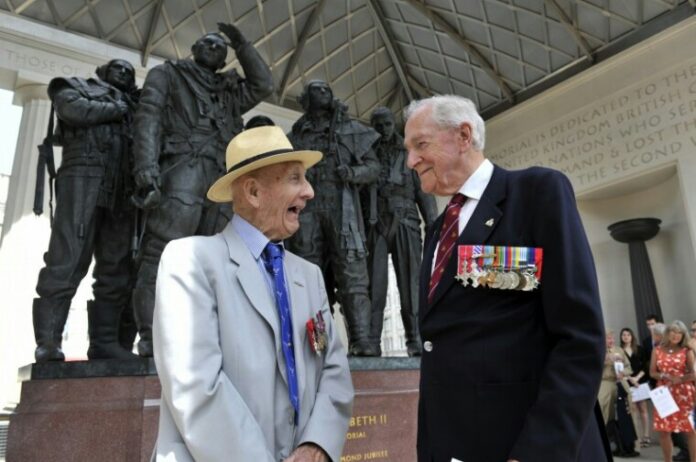 Two elderly men in suits adorned with medals stand smiling in conversation at a military memorial site, their reflections mirroring the strength of the Army pathfinders they once were.