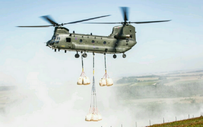 A military helicopter transports large bags suspended below it over a misty landscape.