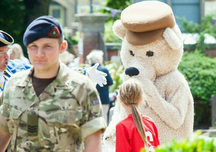 In an outdoor setting, a person dressed in a bear costume entertains children as military personnel in uniform, perhaps on a resettlement assignment, walks past.