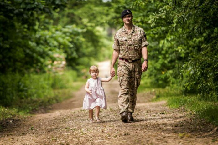 A soldier in camouflage uniform and beret holds hands with a young child in a pink dress, symbolizing hope on their path to resettlement. Walking on a dirt path surrounded by green trees, the serene environment contrasts with the structured life of military duty.