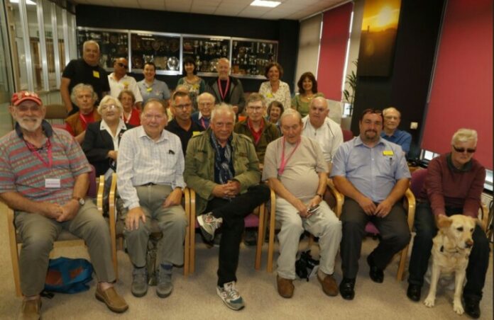 A group of people, mostly older adults including a few veterans, is seated and standing in a room. Some wear lanyards. A dog sits in the front row. Display cases with trophies are visible in the background, adding a sense of pride and achievement to the resettlement gathering.