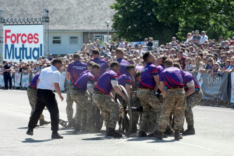 A team of uniformed veterans is pushing a large military cannon in front of a crowd outdoors.