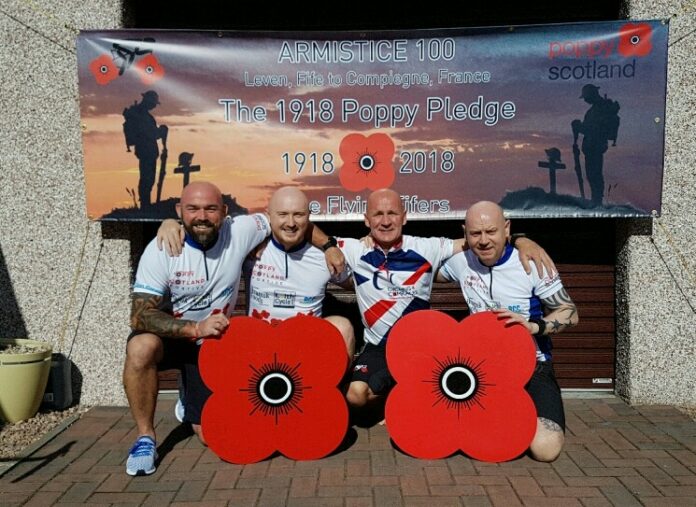 Four men sit in front of a banner for the 1918 Poppy Pledge, holding large poppy symbols. The banner commemorates Armistice 100, featuring poppies and military imagery that honor those who served bravely in the army.