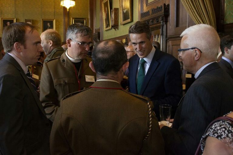 A group of five men in suits and military uniforms, including a seasoned veteran, engage in conversation in an ornate room with portraits on the walls.