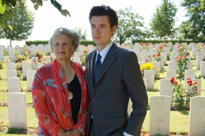 An older woman and a younger man, a veteran freshly resettled, stand together in a cemetery, surrounded by rows of white headstones and rose bushes.