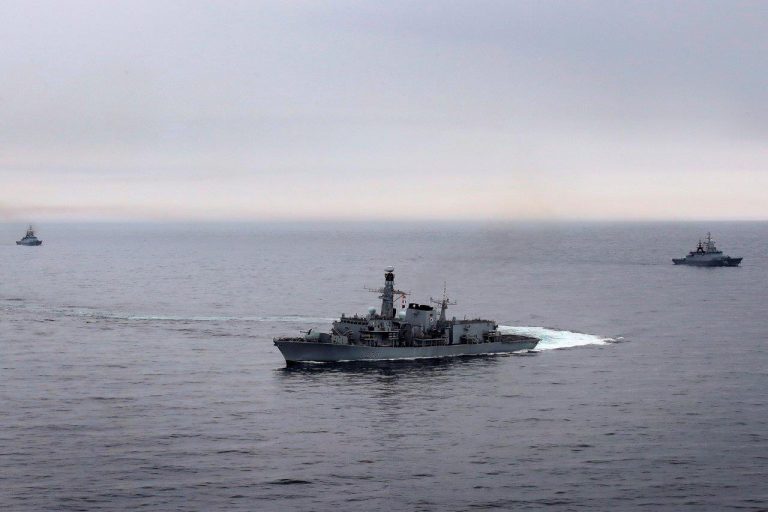 Three naval ships, guided by a seasoned veteran's expertise, maneuver across a calm sea under an overcast sky.