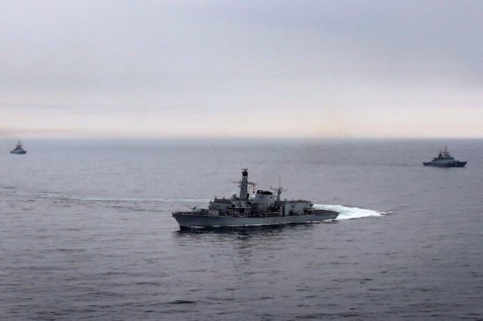 Three naval ships, guided by a seasoned veteran's expertise, maneuver across a calm sea under an overcast sky.