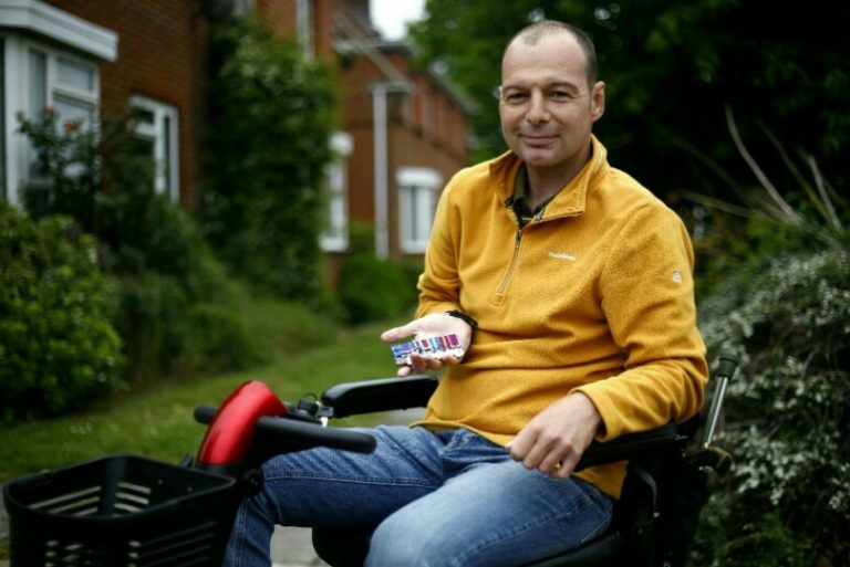 A veteran in a yellow jacket sits confidently in a mobility scooter, holding a small object, with houses and greenery providing a serene backdrop.