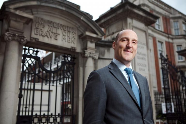 A man in a suit stands in front of a historic building entrance, where an engraved message honors the brave pathfinders of the Royal Navy.
