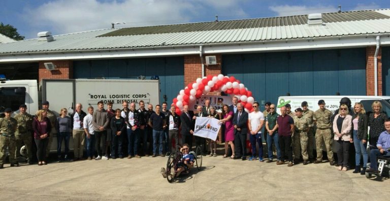 A group of people, including some in Royal Navy uniforms, stand outside a building. At the forefront is a person in a wheelchair, set against a backdrop of red and white balloons.