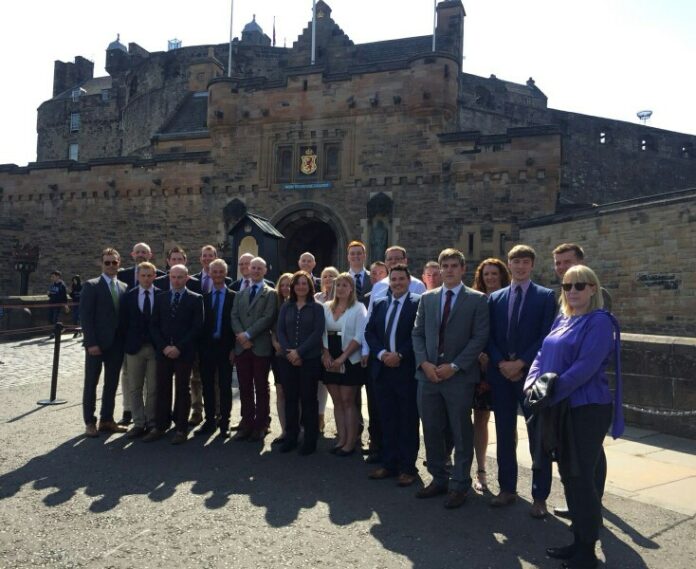 A group of people, including military personnel, stands in front of an old stone building with towers and a large archway entrance on a sunny day.