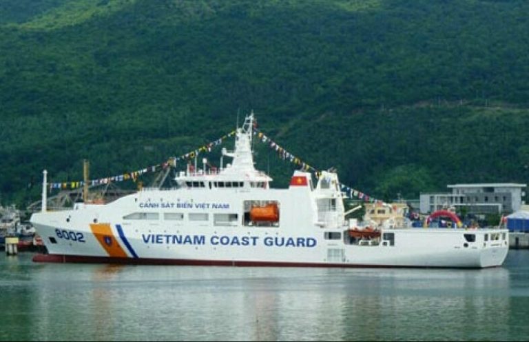 A Vietnam Coast Guard ship, decked with festive flags, is docked in the harbor against a mountainous backdrop. The scene evokes a sense of resettlement, reminiscent of an army veteran finding peace amidst nature's splendor.