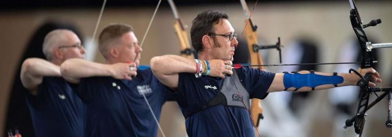 Three archers in blue shirts, reminiscent of Royal Navy precision, aim arrows with their bows in a line at an indoor range.