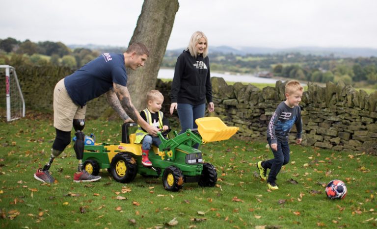 A veteran with a prosthetic leg, a woman, and two children enjoy playing with a toy tractor and a ball in their resettlement home's grassy yard, surrounded by trees and a stone wall.