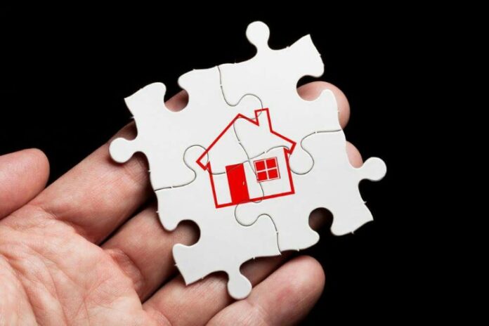 A hand holds a white puzzle piece with a red house illustration, symbolizing the intricate journey of resettlement, against a black background.