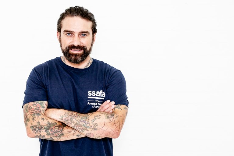 A bearded veteran with tattoos, wearing a navy blue T-shirt, stands with his arms crossed against a white background.