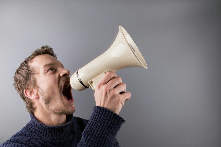 A man in a dark sweater is shouting into a cream-colored megaphone against a gray background, as if commanding an army.