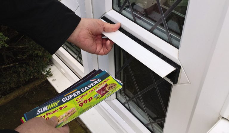 A person is stealthily inserting Subway flyers, labeled "Super Savers," into a letterbox on a door with diamond-patterned glass panes, much like a pathfinder on a mission to deliver delicious deals.