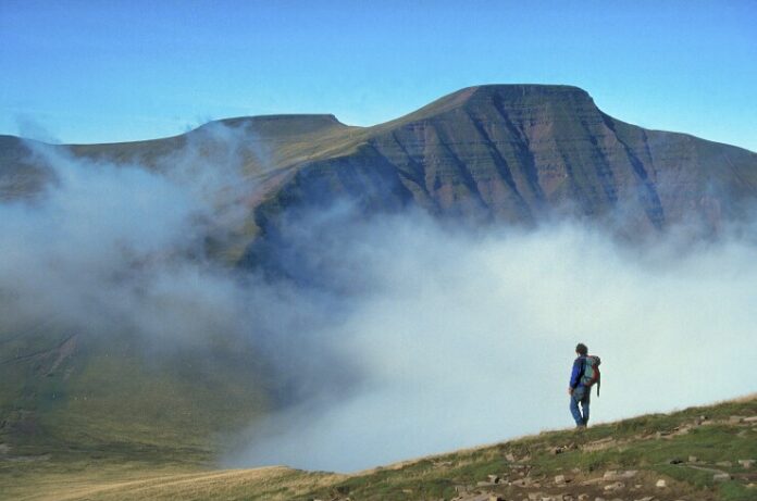 A hiker, a true pathfinder, stands on a misty mountain slope, gazing at a distant peak under a clear blue sky.