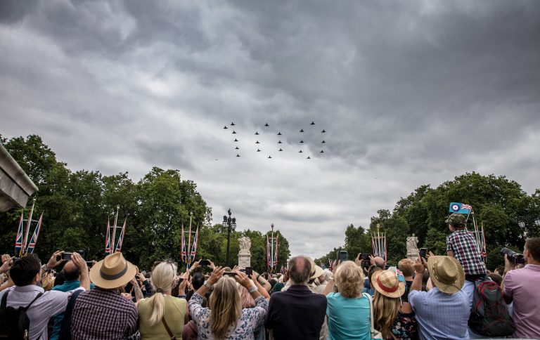 A crowd watches as a formation of military jets soars overhead under cloudy skies. Many people, captivated by the display, are capturing the moment with their phones. Flags and greenery adorn the background, adding to the sense of pride and patriotism.