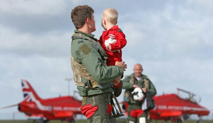 A person in a flight suit holds a child dressed in red. Another individual, possibly a military veteran, stands nearby in flight gear. Two red jets adorned with the Union Jack are in the background.