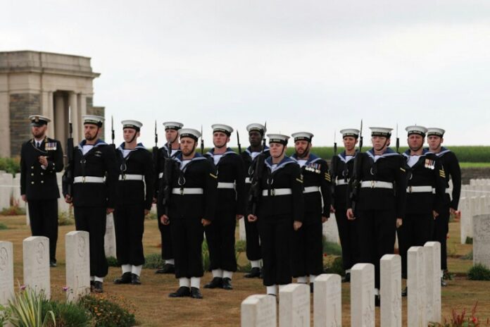 A group of Royal Navy sailors in formal uniforms stands in formation among white gravestones in a cemetery, honoring the fallen veterans.