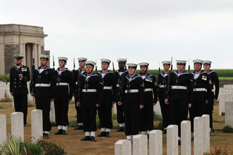 A group of Royal Navy sailors in formal uniforms stands in formation among white gravestones in a cemetery, honoring the fallen veterans.
