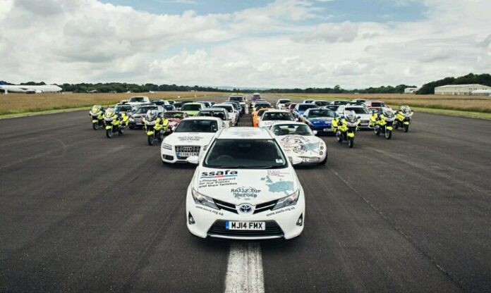 A large convoy of various vehicles, including motorcycles and police cars, is parked on a runway. The leading car, bearing military decals and text on the hood, stands out in this assembly.