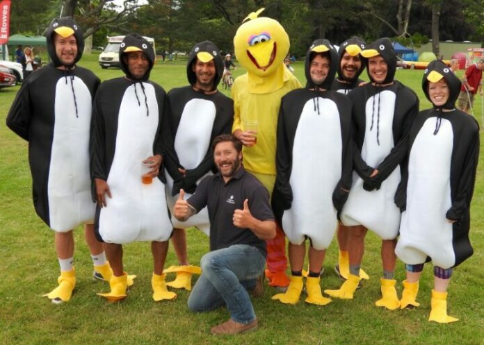A group of people in penguin and bird costumes stand on grass like a whimsical army, with one person kneeling in front, giving a thumbs up.
