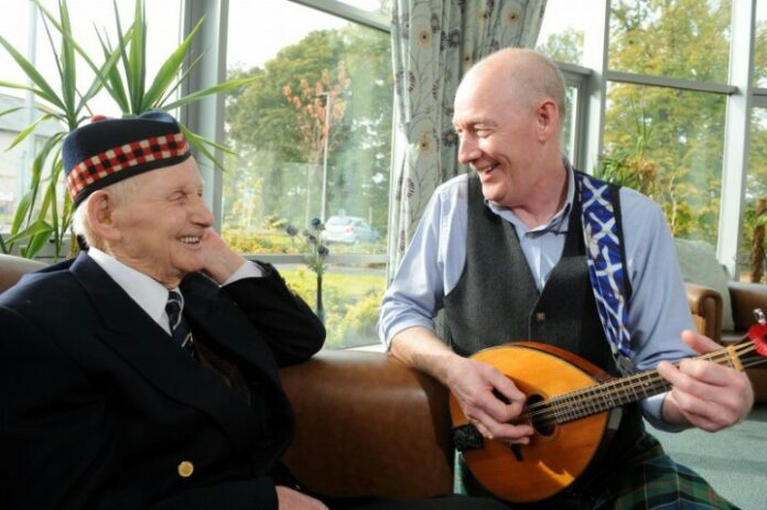 Two men in traditional attire, one elderly veteran seated and smiling, the other standing and playing a stringed instrument in a room with large windows and lush plants.