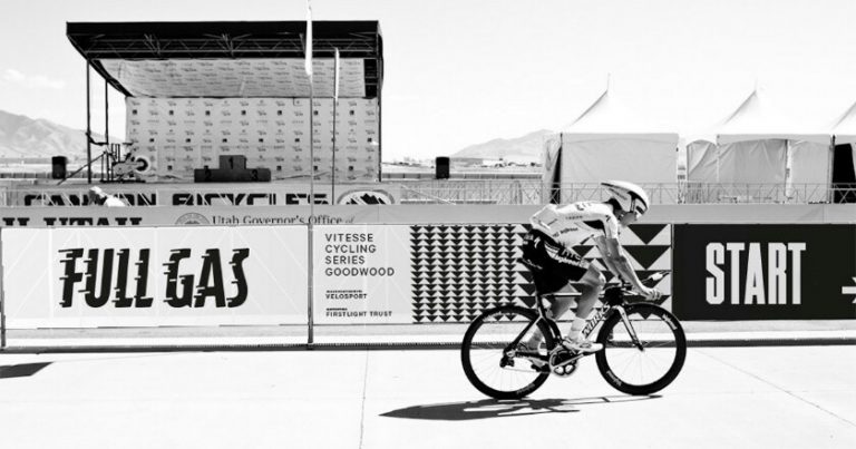 A cyclist smoothly rides past a "START" sign on the track, embodying the spirit of a pathfinder, with a bustling stage and tents in the background.