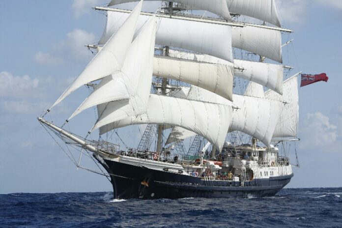 A large, multi-masted sailboat, reminiscent of a Royal Navy veteran, is navigating through the ocean with its white sails billowing under the expansive blue sky.
