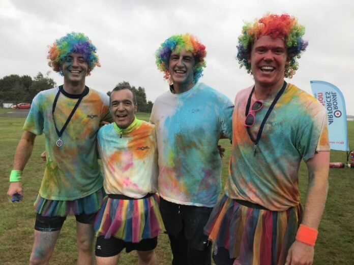 Four people wearing rainbow wigs and colorful outfits pose and smile outdoors, evoking the vibrant camaraderie reminiscent of a Royal Navy celebration.