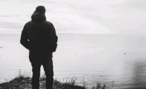 A veteran in a jacket stands on a grassy ledge, gazing over a calm body of water under a cloudy sky, embodying the spirit of a pathfinder.